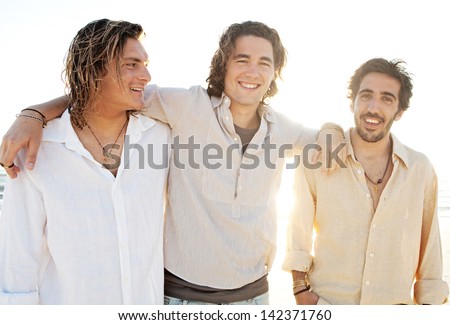 Three young men friends hanging around on vacation together with their arms around each other at sunset while on a beach having fun during the summer.