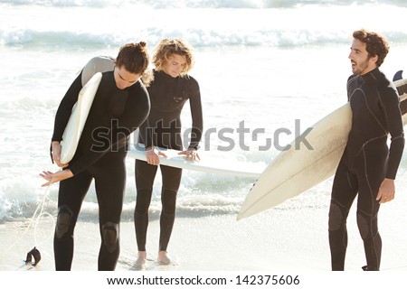 Three sport surfer friends standing together on a white sand beach carrying their surfing boards near the shore during a sunny golden day on vacation, getting ready for a surf session.