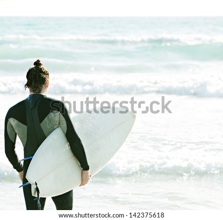 Rear view of a young surfer sport man carrying his surfing board under his arm and walking into the sea with waves during a sunny day on vacation.