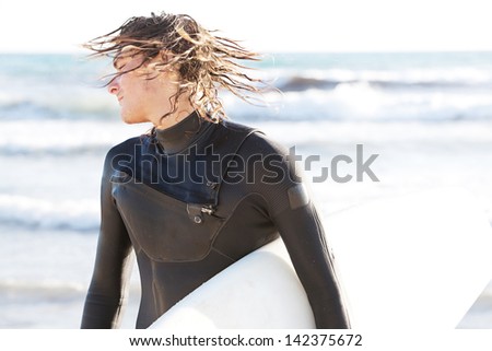 Attractive young surfer shaking the water off his hair on the shore after a surfing session in the sea, carrying his surfing board under his arm while doing sport on vacation.