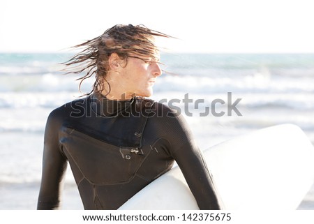 Attractive young surfer shaking the water off his hair on the shore after a surfing session in the sea, carrying his surfing board under his arm while doing sport on vacation.