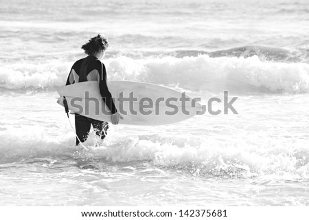 Black and white rear view of a confident young surfer man carrying his surfing board under his arm and walking into the sea with large waves during a sunny day on vacation.