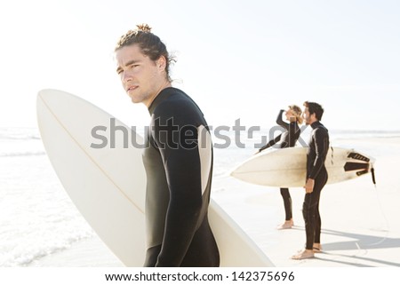 Three surfing sports men standing together on a white sand beach shore, looking at sea and getting ready to surf with one man turning to look at the camera during a sunny day.