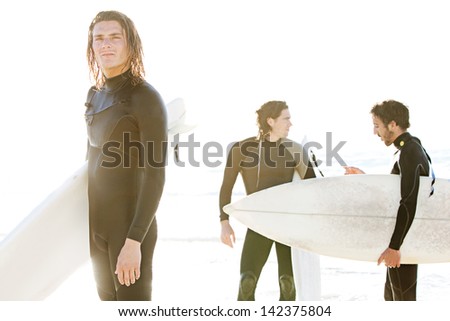 Team of three surfer men standing on a beach with their surfing boards after a surfing session, with the sunset and a golden glare light, while on vacation together.