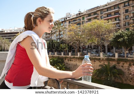 Young woman taking a break from exercising in the city, holding a blue plastic bottle of mineral water and with a white towel over her shoulder during a sunny morning.