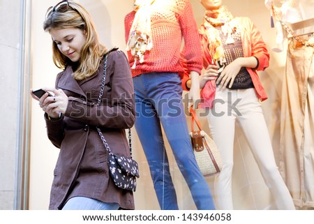 Young stylish teenager woman using a smartphone device while visiting the city, leaning on a fashion store shop window while shopping for clothes.