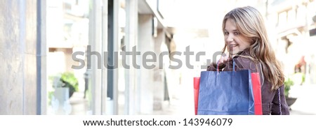 Panoramic view of a young woman walking and shopping in the city, turning to smile at camera while carrying paper bags over her shoulder, joyful and smiling.