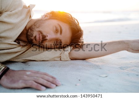 Close up portrait of a young attractive man lying down and relaxing on a white sand beach with the sunset behind him with flare and warm flooding light, on vacation.