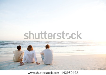 Rear view of three friends men sitting together on a white sand beach with the sun setting behind them with warm orange light, during their vacation in an idyllic nature scene destination.