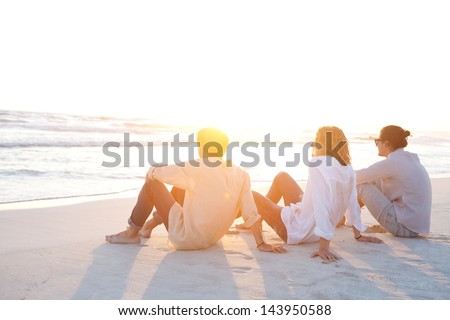 Rear View Of Three Friends Men Sitting Together On A White Sand Beach With The Sun Setting Behind Them With Warm Orange Light, During Their Vacation In An Idyllic Nature Scene Destination.