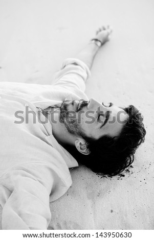 Black and white image of a young attractive man relaxing laying down on the shore of a white sand beach with his arms outstretched and his eyes closed.