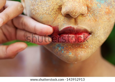 Close up beauty portrait of a young girl wearing red glossy lipstick, make up color powder and glitter on her face with her hand rubbing off her lipstick and staining her face. Texture detail.