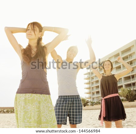 Group of three friends dancing, listening to music and having fun on an urban beach in the city with their arms up against the sunshine filtering trough them.