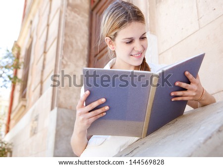 Teenager student girl reading her notebook while leaning on the banister of an old stone university college building, focused and smiling.