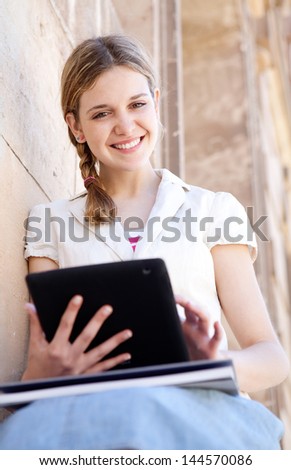 Teenager student using a touch screen digital pad to study while sitting down on the side of an old stone college university building, smiling.