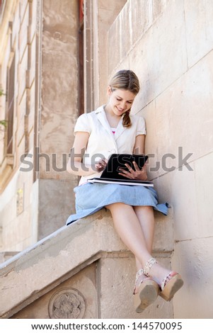Teenager student using a touch screen digital pad to study while sitting down on the banister of an old stone college university building, smiling.