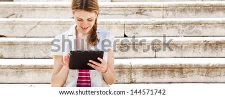 Panoramic portrait of a teenager student girl using with her digital tablet to go on-line while sitting down on a university campus entrance stone steps, smiling during  a sunny day.