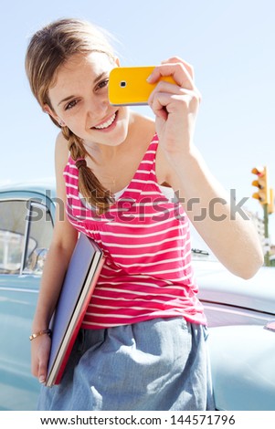 Portrait of a teenager girl using with her smartphone to take photos while leaning on a classic car in the city during a sunny day with a blue sky, smiling.