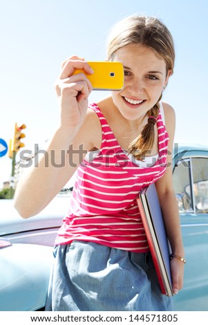 Portrait of a teenager girl using with her smartphone to take photos while leaning on a classic car in the city during a sunny day with a blue sky, smiling.