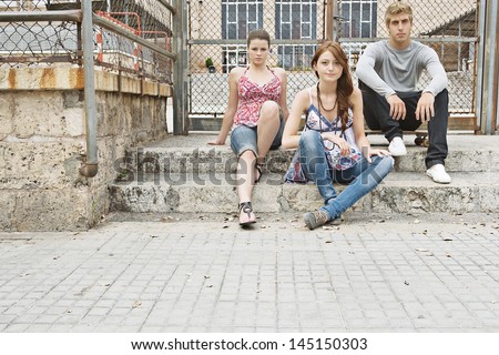 Group of three attractive teenagers sitting down on the stone steps of a college entrance, relaxing and being fashionable, the young man sitting on a skateboard.
