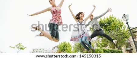 Panoramic view of a group of three friends jumping up in the air together while visiting an urban park in the city, having fun and enjoying the energy during a summer day on vacation.