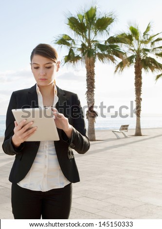 Young professional office businesswoman standing in a clean and open space near the sea, holding and using a digital technology tablet with a touch screen against a blue sky.