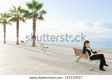 Young office business woman sitting on a wooden bench by the sea, holding an mp4 player and listening to music while relaxing with a blue sky and palm trees during a sunny day.