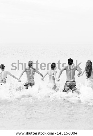 Black and white view of a group of five mixed friends holding hands and running into the sea water together, being spontaneous and having fun while on a summer vacation on a beach.