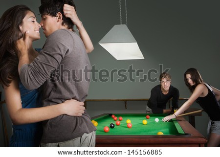 Two young couples playing pool together in a bar while having a night out in town with one couple hugging and kissing with passion, interior.