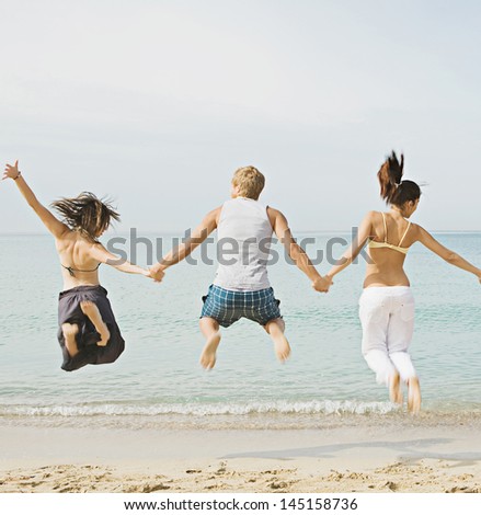 Group of three friends holding hands and jumping at once on the shore of a golden sand beach against a blue sea and sky, expressing energy, fun and joy.