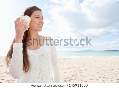 Young attractive healthy woman on a white sand beach holding a sea shell against her ear and listening to the sound of waves smiling, during a vacation trip away.