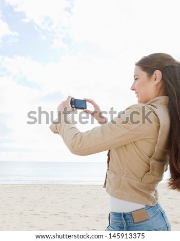 Side view of a young woman standing on a white sand beach, taking pictures of the scenery during her vacation with her digital photo camera.