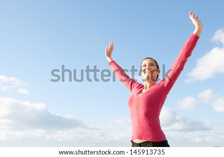 Attractive young woman standing with her arms outstretched up in the air above her head, joyfully smiling against an intense blue sky during a sunny day.