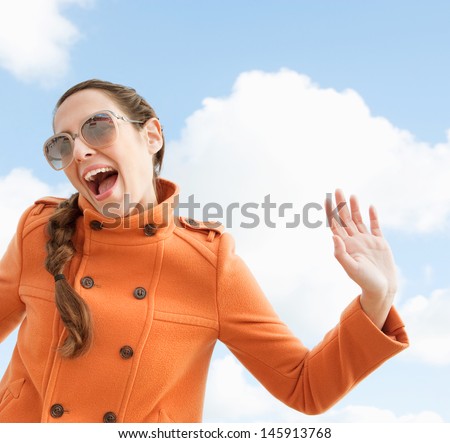 Attractive quirky individual woman shouting and holding her arms in the air with a humorous fun expression against a blue sky with clouds, outdoors.