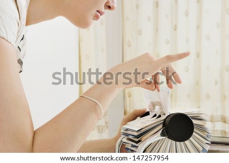 Close up profile view of a young business woman leaning forward on her office desk and picking a business card contacts details from her roller deck database, interior.
