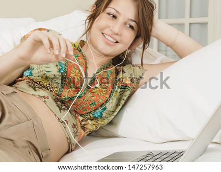 Beautiful young woman lounging on her sofa at home and listening to music with her laptop computer and headphones, smiling and enjoying the moment.