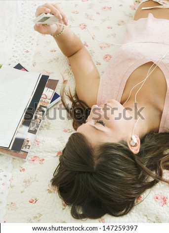 Over head view of a beautiful teenager relaxing and laying down on her bed in her bedroom at home, using an mp3 player and headphones to listen to music.
