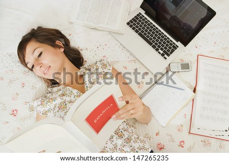 Over head view of a teenager student of languages doing her homework, laying on her bed in her bedroom with a laptop computer and cell phone, reading an open Spanish book.