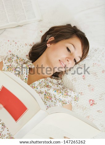 Over head view of a teenager student of languages doing her homework, laying on her bed in her bedroom with a laptop computer and cell phone, reading an open book.