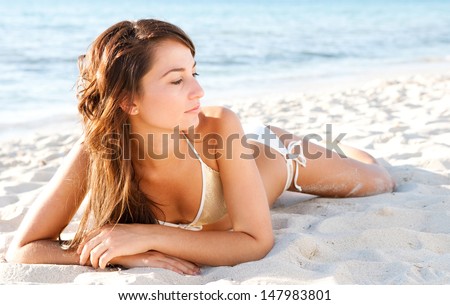 Beautiful young woman laying down on a white sand beach on vacation, sunbathing and being thoughtful while relaxing wearing a sequins bikini during sunset.