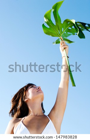Portrait view of a beautiful young woman holding a large exotic green leave up in the air with her arm raised against an intense blue sky. Outdoor nature space.