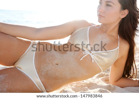 Close up beauty portrait of a young attractive woman laying down and relaxing lounging on a white sand beach shore wearing a gold sequins bikini against the sunset, being sexy.