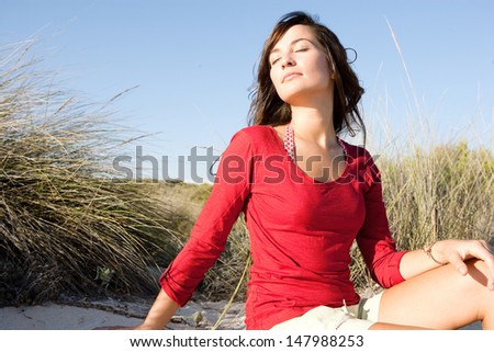 Portrait of a beautiful young woman breathing fresh air while sitting on a beach sand dunes with grass, leaning her head back and enjoying the breeze on vacation.