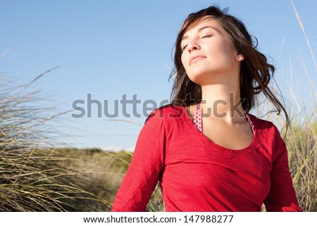 Close up portrait of a beautiful young woman breathing fresh air while sitting on a beach sand dunes with her eyes closed, leaning her head back and enjoying the breeze on vacation.
