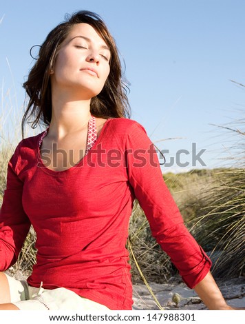 Portrait Of A Beautiful Young Woman Breathing Fresh Air While Sitting On A Beach Sand Dunes With Grass, Leaning Her Head Back And Enjoying The Breeze On Vacation.