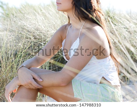 Close up middle section view of an attractive young woman relaxing on the sun dunes of a beach, sitting down and being thoughtful during a sunny day on vacation.