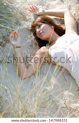 Over head close up portrait of a young beautiful woman laying down on a beach sand dunes, relaxing and feeling the vegetation during a sunny day on holiday.