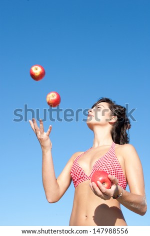 Attractive young woman juggling red apples and wearing a matching red bikini while standing on a beach against an intense blue sky during a sunny day on vacations.
