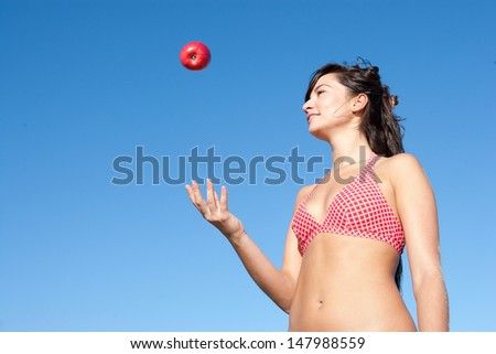 Attractive young woman throwing a red apple up in the air while wearing a matching red bikini and standing on a beach against an intense blue sky during a sunny day on vacation.