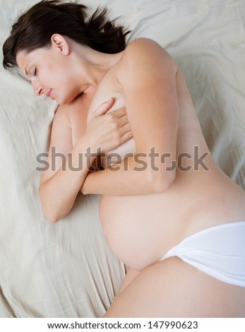 Over head profile view of a young pregnant woman and mum to be sleeping on a bed at home, relaxing and resting with a bare top and wearing white shorts, interior.